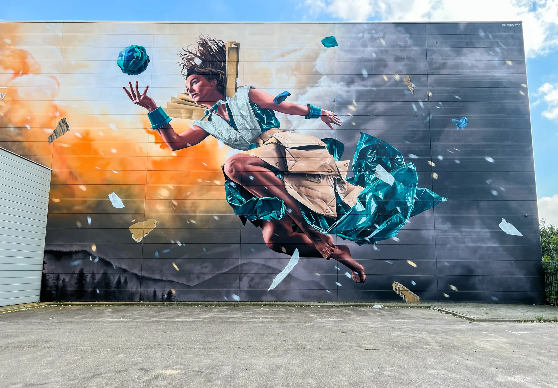 Photo d’une œuvre d’art urbaine sur laquelle on peut voir une femme géante en robe. Derrière elle, on peut voir des nuages, des flammes et des montagnes.  