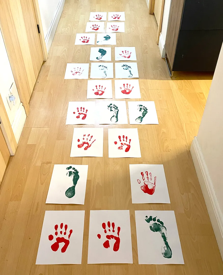 Photographie d’un jeu des pieds et des mains. Des feuilles de papier sont arrangées au sol, elles ont des empreintes de pieds et de mains faites en peinture. 