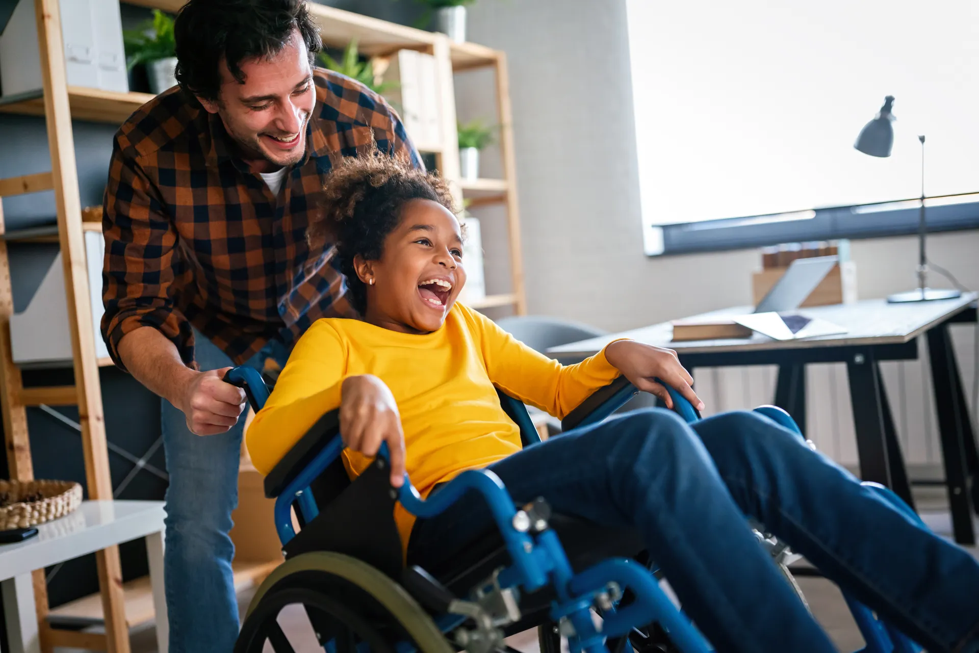 On peut voir un homme qui pousse un enfant en fauteuil roulant. L’homme semble rire et l’enfant affiche un large sourire.